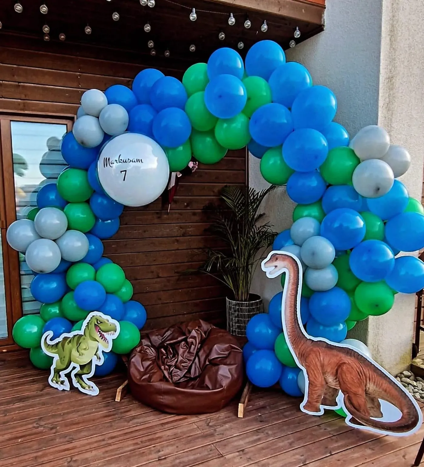 Zila, zaļa un pelēka balonu arka ar dinozauru figūrām un vārda balonu